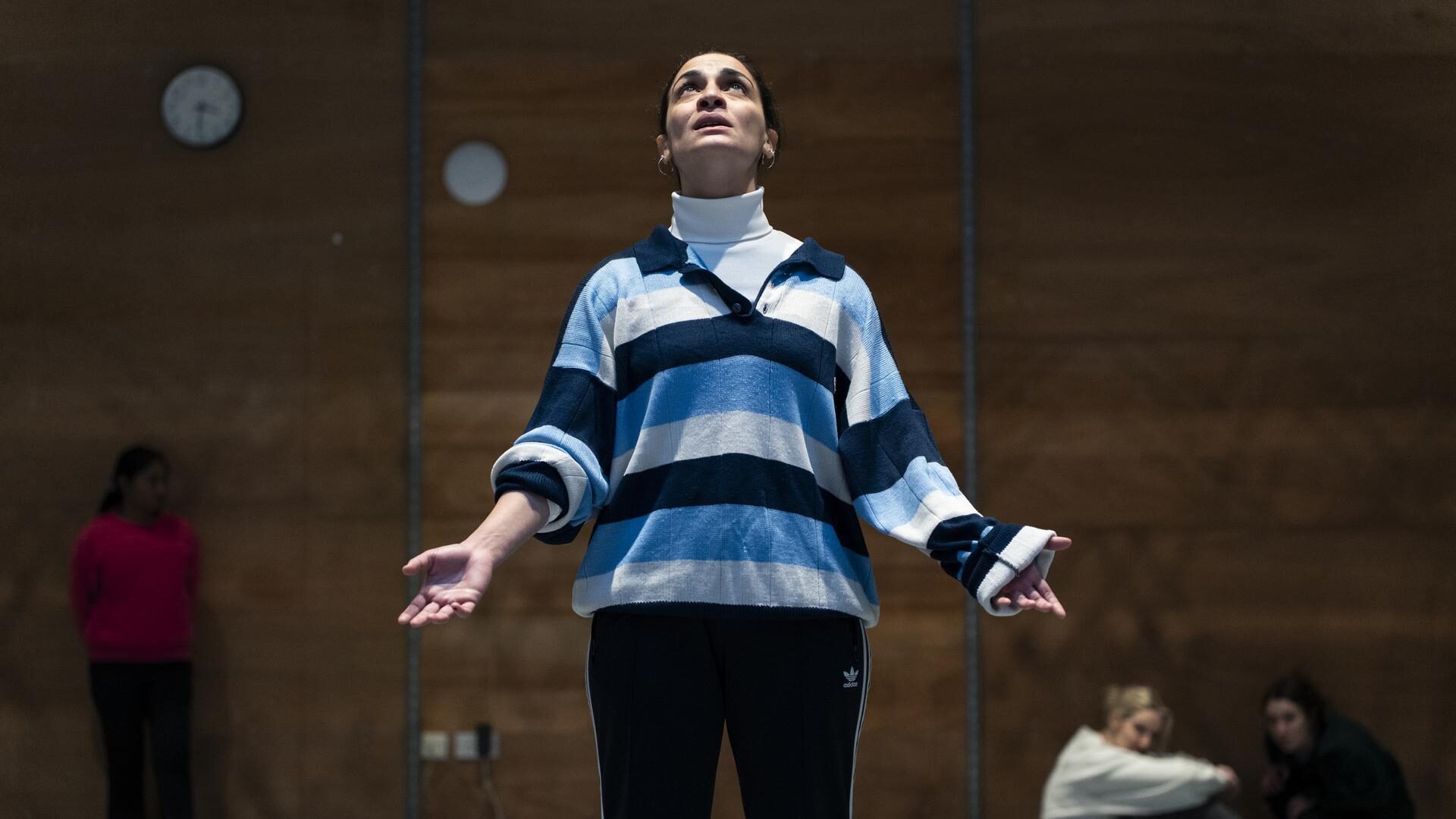 Woman in striped jumper stands with arms outstretched, looking upward during a rehearsal, as others sit nearby